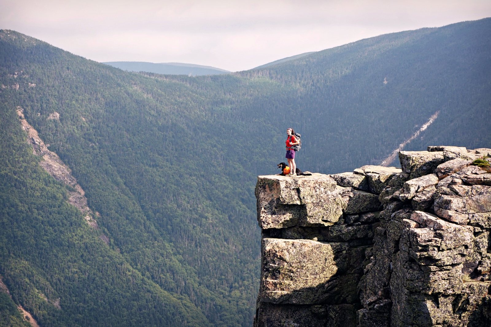 A hiker and her dog stand on the edge of Bondcliff above the Pemigewasset Wilderness